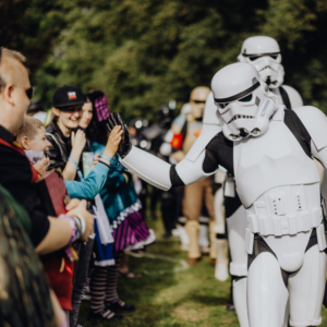 Stormtrooper interagiert mit begeisterten Fans beim Elbenwald Festival, während Teilnehmer in kreativen Kostümen eine lebhafte Atmosphäre genießen. Die Veranstaltung kombiniert Musik und Nerd-Kultur in einem einzigartigen Erlebnis für alle Altersgruppen.