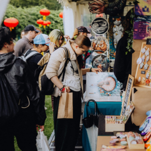 Besucherinnen und Besucher des Elbenwald Festivals erkunden einen Stand mit Kunstwerken und Merchandise. Im Vordergrund betrachtet eine junge Frau ein Plakat, während andere Interessierte in der Warteschlange stehen, um einzukaufen.