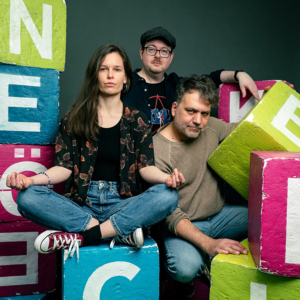 Drei Personen posieren kreativ zwischen bunten Blöcken mit Aufschriften, die die Atmosphäre des Elbenwald Festivals widerspiegeln. Der Hintergrund ist schlicht, um die lebendigen Farben der Blöcke und die Ausdrucksformen der Teilnehmer hervorzuheben.