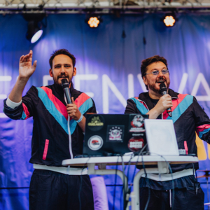 Zwei Moderatoren auf der Bühne des Elbenwald Festivals, die mit Mikrofonen in der Hand das Publikum unterhalten. Sie tragen auffällige Outfits und stehen vor einem Tisch mit technischen Geräten und Notizen, während die Bühnenbeleuchtung im Hintergrund leuchtet.