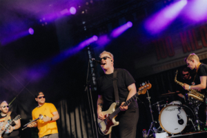 100 Kilo Herz Musiker auf der Bühne des Elbenwald Festivals, umgeben von bunten Lichtern. Die Band spielt energetische Musik, während das Publikum begeistert zuschaut. Eine lebendige Atmosphäre, die die Verbindung von Musik und Nerd-Kultur feiert.