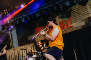 100 Kilo Herz Musiker spielt Trompete auf der Bühne beim Elbenwald Festival. Im Hintergrund sind Bandkollegen und bunte Lichter zu sehen, die die lebendige Atmosphäre des Festivals unterstreichen. Die Energie der Veranstaltung zieht die Zuschauer in ihren Bann.