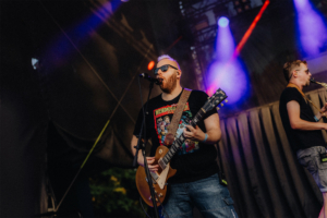 100 Kilo Herz Ein Musiker mit Bart und Sonnenbrille spielt Gitarre auf der Bühne des Elbenwald Festivals. Im Hintergrund ist ein weiterer Musiker mit Saxophon zu sehen, umgeben von bunten Lichtern und einer energiegeladenen Festivalatmosphäre.