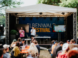 Pen & Paper 1x1 Zwei Moderatoren stehen auf der Bühne des Elbenwald Festivals, während das Publikum aufmerksam zuhört. Im Hintergrund ist ein Bildschirm mit einer Präsentation sichtbar, die verschiedene Themen zeigt. Die Atmosphäre ist lebhaft und kreativ.