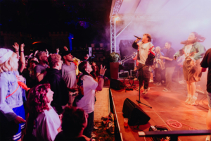 #zweiraumsilke Bühne beim Elbenwald Festival mit einer energiegeladenen Band, die vor begeistertem Publikum auftritt. Die Zuschauer heben die Hände und feiern die Musik in einer bunten, festlichen Atmosphäre. Nebel und Lichter schaffen eine lebendige Stimmung.