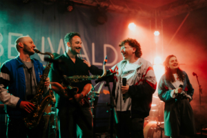 #zweiraumsilke Vier Musiker auf der Bühne des Elbenwald Festivals, lächelnd und interagierend. Zwei spielen Saxophon, während die anderen mit Mikrofonen in der Hand das Publikum unterhalten. Bunte Lichter und eine lebendige Atmosphäre prägen die Szene.