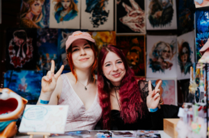 Artist Alley Zwei junge Frauen posieren lächelnd für die Kamera auf dem Elbenwald Festival, umgeben von bunten Kunstwerken und Merchandise. Die Atmosphäre ist lebhaft, mit einer Mischung aus Kreativität und Begeisterung für die Popkultur.