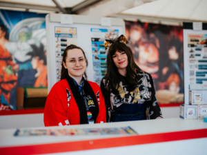 Ichiban Kuji Zwei freundliche Mitarbeiterinnen am Stand des Elbenwald Festivals, umgeben von bunten Hintergrundgrafiken und Merchandise-Artikeln. Die Mitarbeiterinnen tragen kreative Kostüme und lächeln, während sie Besucher willkommen heißen.