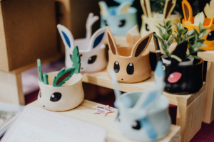 Artist Alley Bunte Blumentöpfe in Form von Pokémon-Charakteren auf einem Holzregal, präsentiert auf dem Elbenwald Festival. Die Töpfe zeigen kreative Designs mit verschiedenen Gesichtern und Ohren, ideal für Pflanzenliebhaber und Fans der Popkultur.