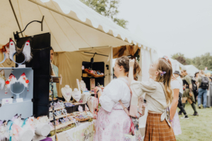 Artist Alley Zwei Besucherinnen des Elbenwald Festivals bewundern handgefertigten Schmuck und Accessoires an einem Stand. Die fröhliche Atmosphäre und die kreative Vielfalt spiegeln die einzigartige Verbindung von Musik und Nerdkultur wider.