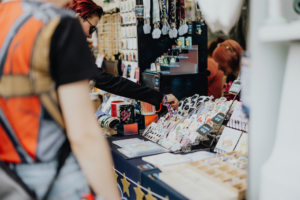 Artist Alley Bunte Merchandise-Stände auf dem Elbenwald Festival, mit Besuchern, die verschiedene kreative Produkte wie Sticker, Tassen und Schlüsselanhänger durchstöbern. Die Atmosphäre ist lebhaft und einladend für Fans von Popkultur und Musik.