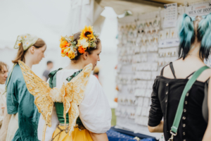 Artist Alley Besucherinnen des Elbenwald Festivals in fantasievollen Kostümen, darunter eine mit Schmetterlingsflügeln und Blumenkranz, erkunden einen Stand mit Schmuck und Accessoires. Die Atmosphäre ist lebhaft und kreativ, ideal für Fans von Fantasy und Musik.