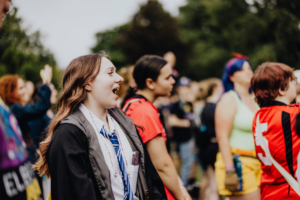 Menschenmenge beim Elbenwald Festival, die begeistert an einem musikalischen Auftritt teilnimmt. Eine junge Frau mit langen Haaren und Krawatte singt laut mit, umgeben von weiteren Festivalbesuchern in bunten Kostümen und T-Shirts.