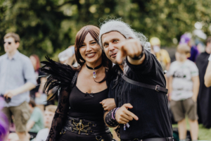 Zwei kostümierte Teilnehmer des Elbenwald Festivals posieren fröhlich für die Kamera. Die Frau trägt ein schwarzes Outfit mit Federn, während der Mann in dunkler Kleidung mit grauen Haaren zeigt. Im Hintergrund sind weitere Festivalbesucher zu sehen.