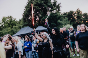 Zwei Frauen in Kostümen halten Lichtschwerter und genießen die Atmosphäre des Elbenwald Festivals. Im Hintergrund sind weitere Festivalbesucher zu sehen, die die Mischung aus Musik und Nerd-Kultur erleben.
