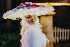 Eine Person mit langen weißen Haaren und einer auffälligen, leuchtenden Pilzkopfbedeckung lächelt in die Kamera. Sie trägt eine weiße Bluse und große Brille, umgeben von einer festlichen Atmosphäre beim Elbenwald Festival.