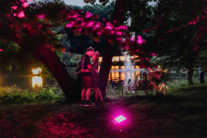 Ein Paar küsst sich romantisch unter einem Baum am Elbenwald Festival, während sanftes, pinkes Licht die Szenerie beleuchtet. Im Hintergrund spiegelt sich der See, was die magische Atmosphäre des Festivals unterstreicht.