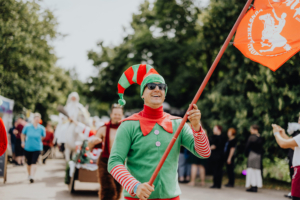 Ompah Ein fröhlicher Elf mit Sonnenbrille und einem roten Fahnenstab, gekleidet in ein grünes und rotes Kostüm, zieht während des Elbenwald Festivals durch die Menge. Im Hintergrund sind weitere festlich gekleidete Teilnehmer und Stände zu sehen.
