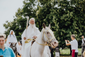 Ompah Ein älterer Mann mit langem, weißem Bart und in weißem Gewand reitet auf einem weißen Pferd, während er einen Stab hält. Im Hintergrund sind weitere Teilnehmer des Elbenwald Festivals zu sehen, die sich in fantasievollen Kostümen bewegen.