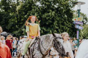 Ein Mädchen mit roten Zöpfen und einem Plüschhund auf der Schulter reitet auf einem gefleckten Pferd während des Elbenwald Festivals. Im Hintergrund sind andere Besucher und Wegweiser zu verschiedenen Attraktionen zu sehen.