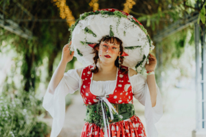 Bunt gekleidete Frau mit großem, dekoriertem Hut und rotem Kleid mit weißen Punkten, steht in einer grünen Umgebung. Ihre Accessoires und das Outfit spiegeln die kreative Atmosphäre des Elbenwald Festivals wider, das Musik und Nerd-Kultur vereint.