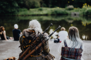 Teilnehmer des Elbenwald Festivals sitzen am Ufer eines Teiches, während ein kostümierter Charakter mit grauen Haaren und Waffen im Hintergrund zu sehen ist. Die Szene vermittelt eine entspannte Atmosphäre inmitten der Natur und der Festival-Teilnehmer.