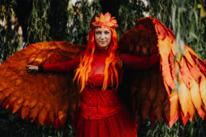 Eine Frau mit leuchtend roten Haaren und einem auffälligen Kostüm mit großen, orangefarbenen Flügeln posiert im Freien. Das Outfit kombiniert Elemente von Fantasy und Natur und spiegelt den kreativen Geist des Elbenwald Festivals wider.