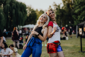 Zwei Frauen in kreativen Kostümen lächeln und posieren fröhlich beim Elbenwald Festival. Im Hintergrund sind andere Festivalbesucher und Stände zu sehen, die die lebendige Atmosphäre des Events widerspiegeln.