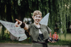 Eine cosplayende Person mit Elfenflügeln und einem freundlichen Lächeln zeigt auf etwas im Hintergrund. Sie hält ein Modell eines Schiffs in der Hand. Die Szene spielt beim Elbenwald Festival, wo Fans von Fantasy und Musik zusammenkommen.