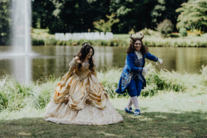 Zwei Cosplayer in aufwendigen Kostümen tanzen am Ufer eines Sees beim Elbenwald Festival. Die Szene wird von einem Wasserfall im Hintergrund und grüner Natur umgeben, was die festliche Atmosphäre des nerdigen Events unterstreicht.