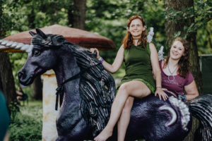 Zwei Frauen posieren auf einem bunten, kunstvollen Einhorn in einem Wald, umgeben von grüner Natur. Die Atmosphäre ist verspielt und fantasievoll, passend zum Elbenwald Festival, das Musik und Nerdkultur vereint.
