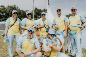 Eine Gruppe von sechs Teilnehmern des Elbenwald Festivals posiert im Freien, gekleidet in gelb-blauen Outfits und Caps mit Pokémon-Motiven. Sie halten Wasserpistolen und lächeln in die Kamera, während Seifenblasen um sie herum schweben.