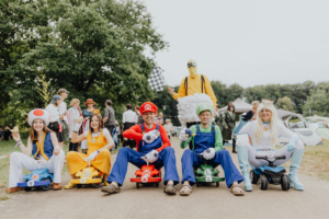 Eine Gruppe von Cosplayern in bunten Kostümen sitzt auf kleinen Fahrzeugen beim Elbenwald Festival. Zu sehen sind Charaktere aus Videospielen, darunter Mario, Luigi und Prinzessin Peach, umgeben von festivaltypischer Atmosphäre und anderen Besuchern. Foto: Gina Wetzler