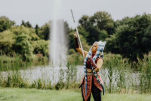 Eine Cosplayerin in einem detailreichen Outfit, inspiriert von einer bekannten Sci-Fi-Figur, posiert am Ufer eines Sees während des Elbenwald Festivals. Im Hintergrund sprudelt ein Wasserfall zwischen üppigem Grün.