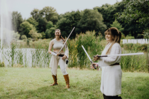 Zwei Cosplayerinnen in fantasievoller Kleidung üben mit Lichtschwertern am Ufer eines Sees während des Elbenwald Festivals. Im Hintergrund ist das Festival-Logo sichtbar, umgeben von grüner Vegetation und einem ruhigen Wasserspiel.