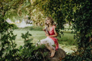 Ein Mädchen in einem pinken Kleid sitzt auf einem Stein, umgeben von grünen Pflanzen. Ihre lockigen Haare fallen sanft über die Schultern, während sie neugierig in die Ferne blickt. Die Szene strahlt eine entspannte Festivalatmosphäre aus.