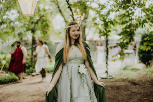 Frau in einem eleganten, grünen Kostüm mit Blumenaccessoire beim Elbenwald Festival, umgeben von einem naturnahen, festlichen Ambiente. Im Hintergrund sind weitere Teilnehmer in Kostümen zu sehen, die die kreative Atmosphäre des Festivals unterstreichen.