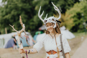 Ein Teilnehmer des Elbenwald Festivals trägt ein kreatives Outfit mit einem Geweih-Hut und einem geschmückten Stock. Im Hintergrund sind Zelte und Festivalbesucher zu sehen, die zur lebhaften Atmosphäre des Events beitragen.