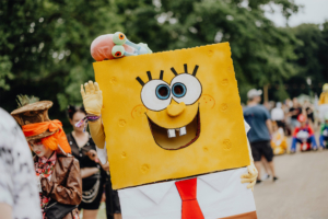 Ein verkleideter Festivalbesucher als SpongeBob Schwammkopf mit einer Plüschfigur von Gary auf dem Kopf, umgeben von einer bunten Menge von Cosplay-Fans. Der Hintergrund zeigt grüne Bäume und andere Teilnehmer des Elbenwald Festivals.