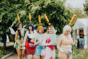 Cosplayer in bunten Kostümen mit Flügeln und auffälligen Accessoires, die beim Elbenwald Festival in einer fröhlichen Gruppe posieren. Die Veranstaltung kombiniert Musik und Nerd-Kultur und bietet eine lebendige Atmosphäre für Fans.