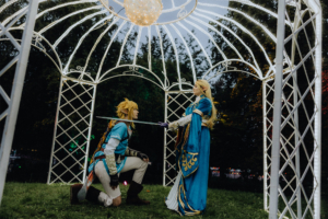 Cosplay-Darstellung von Link und Zelda im festlich beleuchteten Pavillon beim Elbenwald Festival. Link kniet und hält ein Schwert, während Zelda ihn mit einem majestätischen Blick ansieht. Die Szene vermittelt eine magische Atmosphäre und feiert die Fantasiewelt.