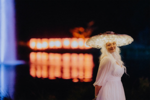 Frau mit leuchtendem Hut und langen, weißen Haaren, trägt ein elegantes, weißes Kleid. Der Hintergrund zeigt eine farbenfrohe Lichtinstallation am Wasser, die die festliche Atmosphäre des Elbenwald Festivals unterstreicht.