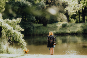 Ein Cosplayer mit blonden Haaren steht am Ufer eines ruhigen Teichs, trägt ein auffälliges schwarzes Cape und hält ein Schwert. Die Szenerie ist von üppigem Grün umgeben, was die Atmosphäre des Elbenwald Festivals unterstreicht.