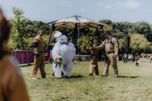 Cosplay auf dem Elbenwald Festival: Zwei Ghostbusters in Kostümen interagieren mit einem aufblasbaren Marshmallow-Man, während im Hintergrund die Festivalbühne und andere Besucher zu sehen sind. Ein lebhaftes Beispiel für die kreative Atmosphäre des Events.