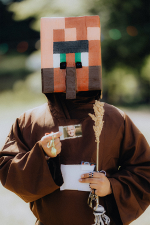 Ein Teilnehmer des Elbenwald Festivals trägt ein kreatives Kostüm mit einem großen, bunten Kopf und brauner Robe. In der Hand hält er ein Namensschild und eine Pflanze, während er in die Kamera lächelt. Hintergrund mit verschwommenen Lichtern.