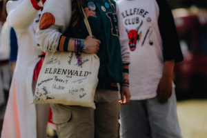 Eine Person trägt eine signierte Elbenwald-Tasche beim Elbenwald Festival, umgeben von anderen Festivalbesuchern in kreativen Outfits. Die Tasche ist mit verschiedenen Namen und Sprüchen verziert, was die Gemeinschaft und das kreative Flair des Events zeigt.
