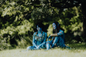 Zwei Cosplayerinnen im Elbenwald Festival, verkleidet als Na'vi-Charaktere aus dem Film "Avatar". Sie sitzen im Gras, umgeben von grüner Natur, und zeigen detaillierte Kostüme mit blauer Farbe und kunstvollen Accessoires.