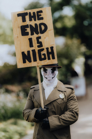 Ein verkleideter Teilnehmer des Elbenwald Festivals hält ein Schild mit der Aufschrift „THE END IS NIGH“. Er trägt einen Trenchcoat, schwarze Handschuhe und hat sein Gesicht maskiert. Die Szenerie zeigt eine bunte Festivalumgebung im Hintergrund.