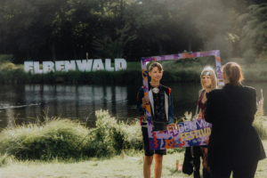 Zwei Jugendliche posieren vor dem Elbenwald Festival Schriftzug am Ufer eines Gewässers. Der Junge hält einen bunten Rahmen mit dem Festival-Logo, während ein Mädchen in Kostüm neben ihm steht. Die Szene vermittelt festliche Stimmung und kreative Ausdrucksformen.