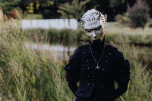 Person mit einer weißen Maske und schwarzer Kleidung steht vor einer grünen Landschaft beim Elbenwald Festival. Im Hintergrund sind die Buchstaben "Elbenwald" sichtbar. Die Atmosphäre kombiniert Elemente von Musik und Fantasy.